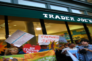 Eine Gruppe von Menschen steht vor einem Geschäft und hält Schilder und Plakate hoch, mit einer Säule und Laternen auf dem Dach im Hintergrund.
