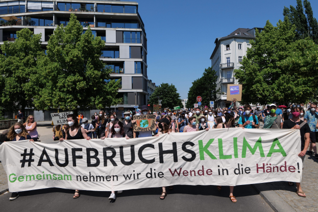 A group of people wearing masks hold a banner reading "Aufbruchsklima" in front of a building with trees and a clear sky.
