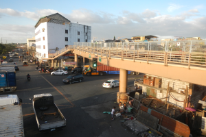 Eine belebte Stadtstraße mit verschiedenen Fahrzeugen, Fußgängern, einer Brücke, Verkehrsampeln, Gebäuden, Bäumen und einem bewölkten Himmel.