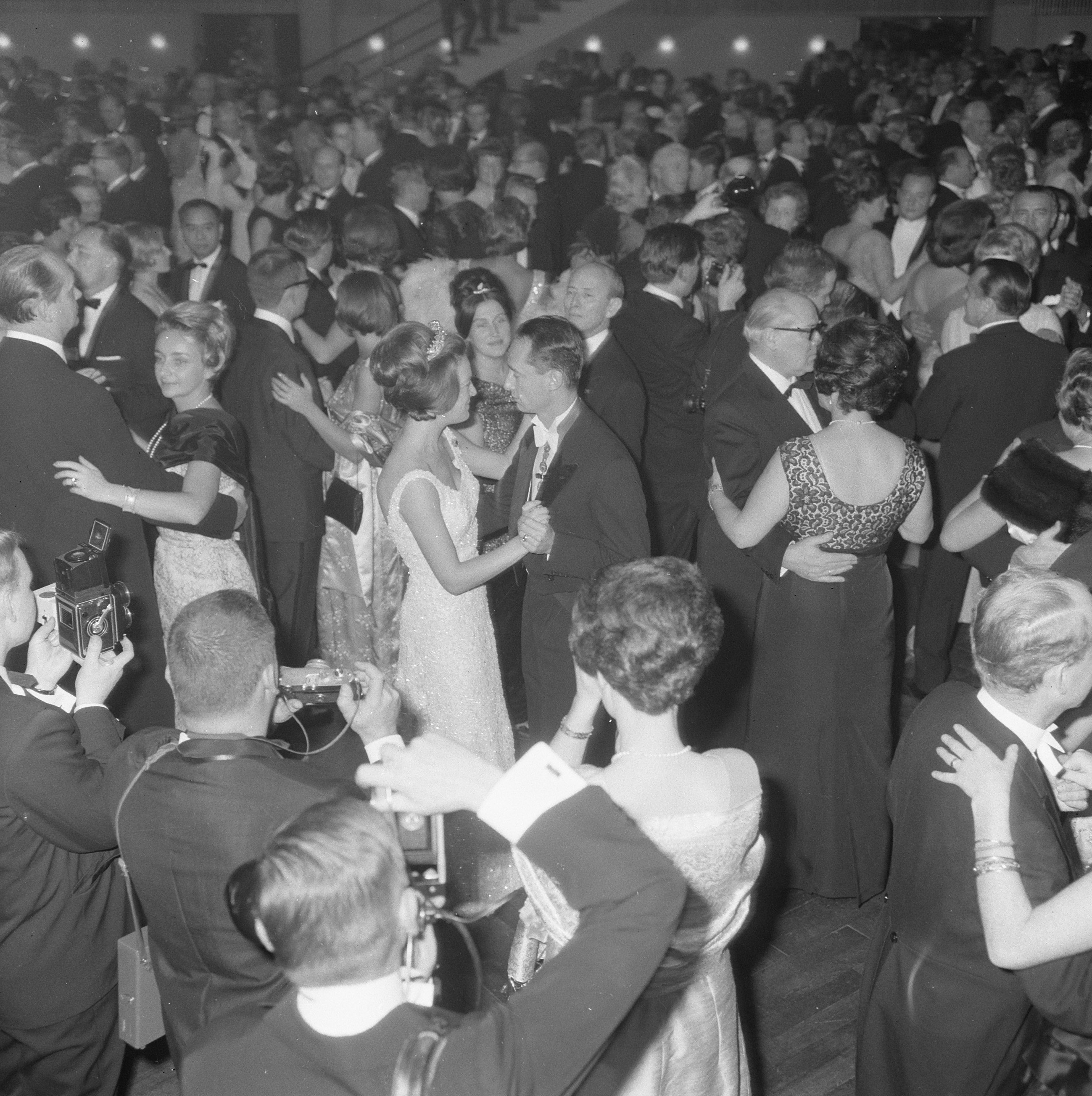 Ein Schwarz-Weiß-Foto einer Gruppe von Menschen auf einer Tanzfläche, einige halten Kameras in den Händen, mit Treppen und Lichtern im Hintergrund.