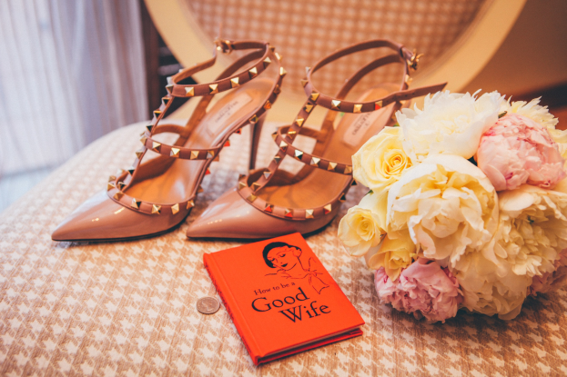 A table display featuring a book titled "How to be a Good Wife," a bouquet of flowers, and a pair of Valentino shoes, set against a curtain backdrop.