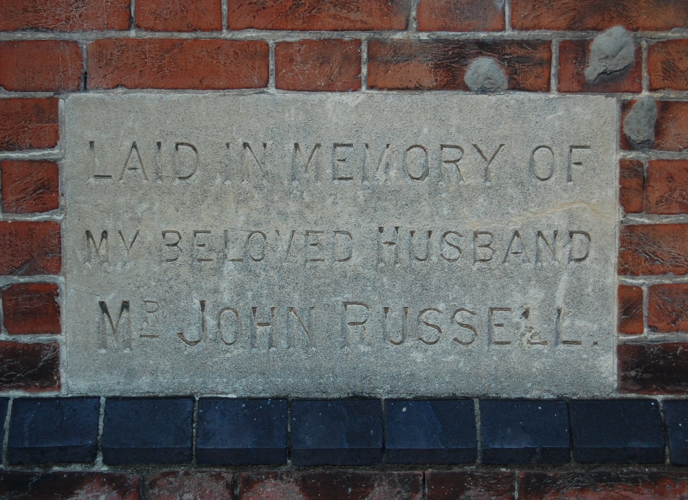 Stone plaque mounted on a brick wall, reading "Laid in Memory of My Beloved Husband".
