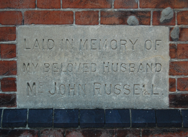Stone plaque mounted on a brick wall, reading "Laid in Memory of My Beloved Husband".