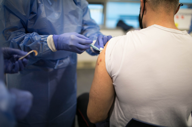 Ein Arzt in einem blauen Kleid und Handschuhen verimpft einem Mann in einem weißen Hemd mit einer Spritze, mit einem unscharfen Hintergrund.