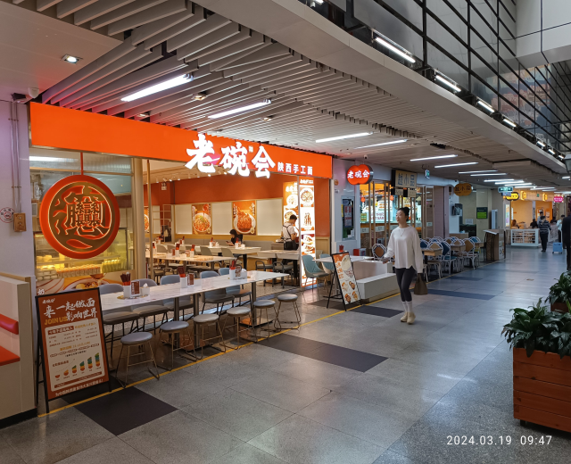 A busy restaurant interior with tables, chairs, and people, featuring ceiling lights, wall-mounted boards, and potted plants on the right.