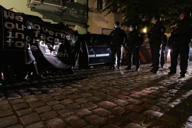 Eine Gruppe von Polizisten in Helmen steht auf einer Kopfsteinpflasterstraße bei Nacht, hält ein Banner, mit Fahrzeugen, Gebäuden, Bäumen und Beleuchtung im Hintergrund.