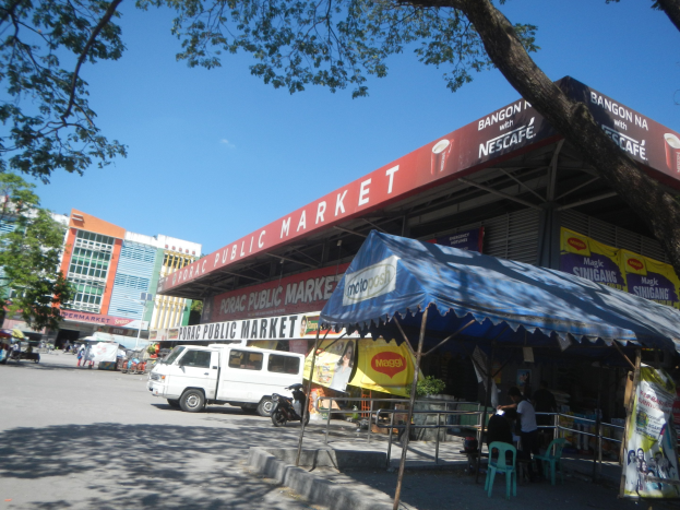Ein belebter Marktplatz nimmt eine Straßenkreuzung ein, mit Gebäuden, Bäumen, Fahrzeugen, Menschen, Stühlen, Tischen, Marktständen, Bannern und einem klaren blauen Himmel.