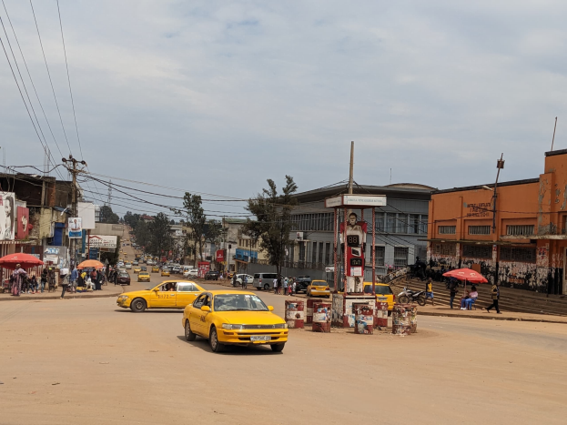 Ein gelbes Taxi fährt auf einer belebten Stadtstraße mit Gebäuden an den Seiten, während Fußgänger mit Regenschirmen auf dem Gehweg unter bewölktem Himmel gehen, mit Strommasten und Kabeln im Hintergrund.