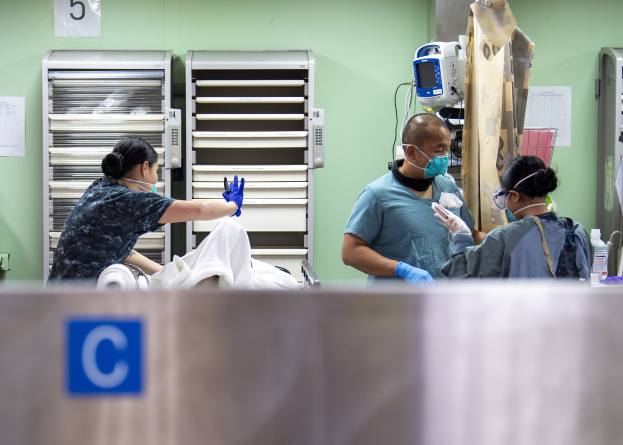 Three healthcare workers in scrubs and masks attend to a patient on a bed in a hospital room, with medical supplies on a table and equipment in the background.