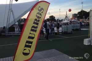 Ein buntes Karnevalszelt mit Zelten, einem zentralen Mast, Menschen, Bäumen, Strommasten, Straßenbahnen auf der Straße und einem eisernen Stand auf der linken Seite unter einem klaren Himmel.