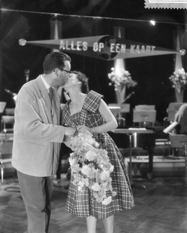 Ein Mann und eine Frau umarmen und küssen sich auf einer Bühne, wobei die Frau einen Blumenstrauß in den Händen hält, umgeben von Stühlen, Blumenvasen und anderen Gegenständen.