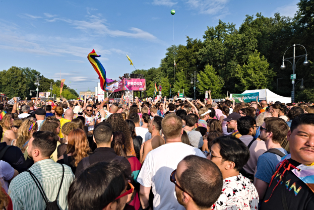 Eine große Menschenmenge versammelt sich vor einem Gebäude, einige halten Fahnen und Banner, mit einem Zelt, Laternenmasten, Bäumen und einem Luftballon im Hintergrund, feiernd die LGBT-Gemeinschaft.