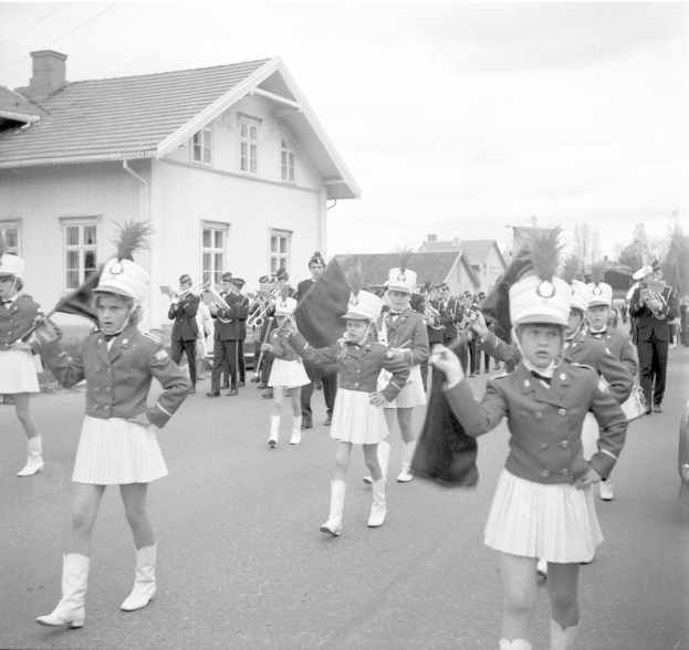 Gruppe junger Mädchen in Uniform, die eine Straße entlangmarschieren, mit Mützen und Fahnen, im Hintergrund Gebäude, Bäume und ein Fahrzeug.