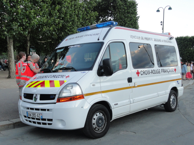 Ambulanz am Straßenrand mit Gruppe von Menschen in roten Jacken in der Nähe, Bäume und Laternen im Hintergrund bei klarem blauem Himmel.