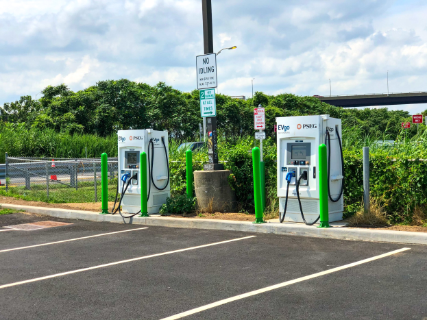 Elektrofahrzeug-Ladestation auf einem Parkplatz mit umgebenen Infrastruktur, Grünflächen und einer Brücke im Hintergrund bei bewölktem Himmel.