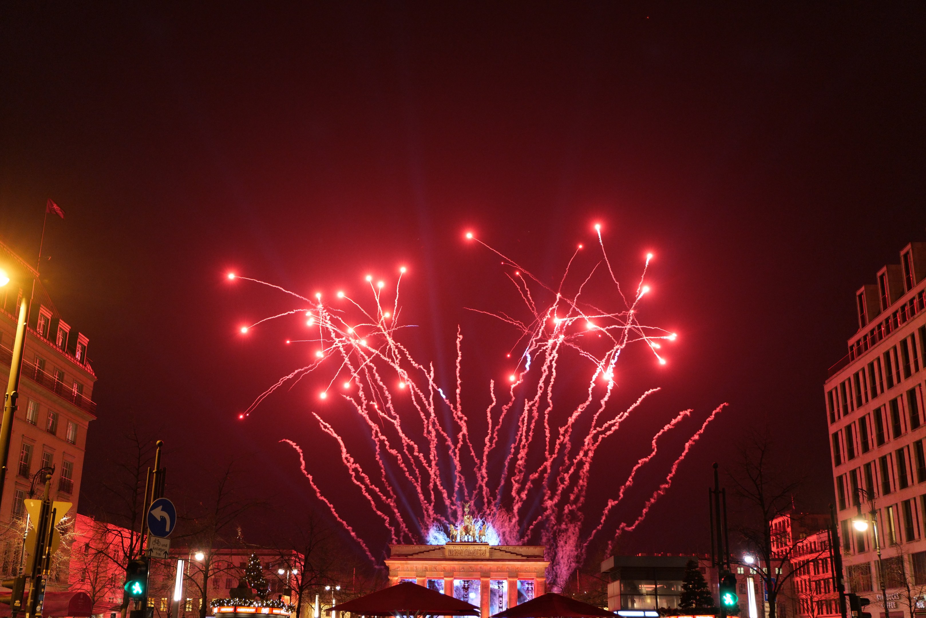 Eine nächtliche Straßenansicht in Berlin am Silvesterabend, mit Gebäuden, Bäumen, Laternenmasten, Verkehrsampeln, Schildern, Zelten, Menschen und einem prächtigen Feuerwerk am Himmel.