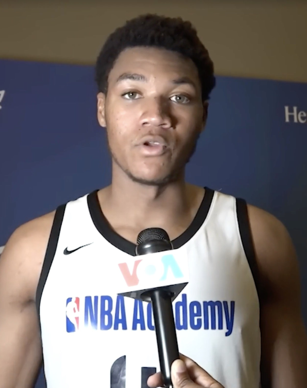 Ein Mann in einem weißen Tanktop hält ein Mikrofon und spricht mit der Medien bei der NBA All-Star-Game 2019, mit einem Banner und einer Wand im Hintergrund.