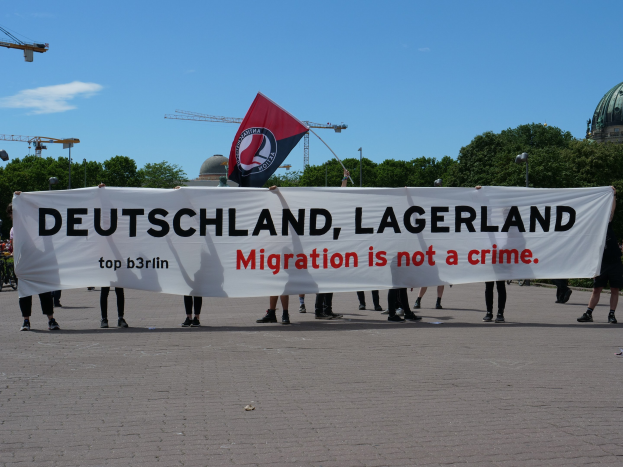 Eine Gruppe von Menschen mit einem Banner "Deutschland, Lagerland Migration ist kein Verbrechen" und einer Flagge, mit Laternenmasten, Lichtern, Bäumen, Kränen, einem Gebäude und einem bewölktem Himmel im Hintergrund.