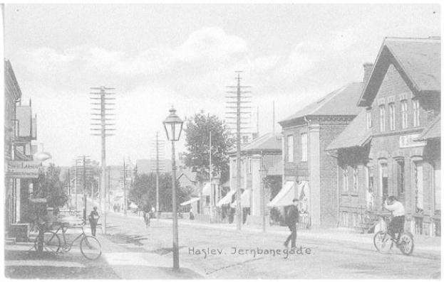 Schwarzes und weißes Foto einer Stadtstraße in Haslev, Jernbangegade, mit Menschen auf Fahrrädern, Strommasten, Straßenlaternen, Bäumen und Gebäuden.