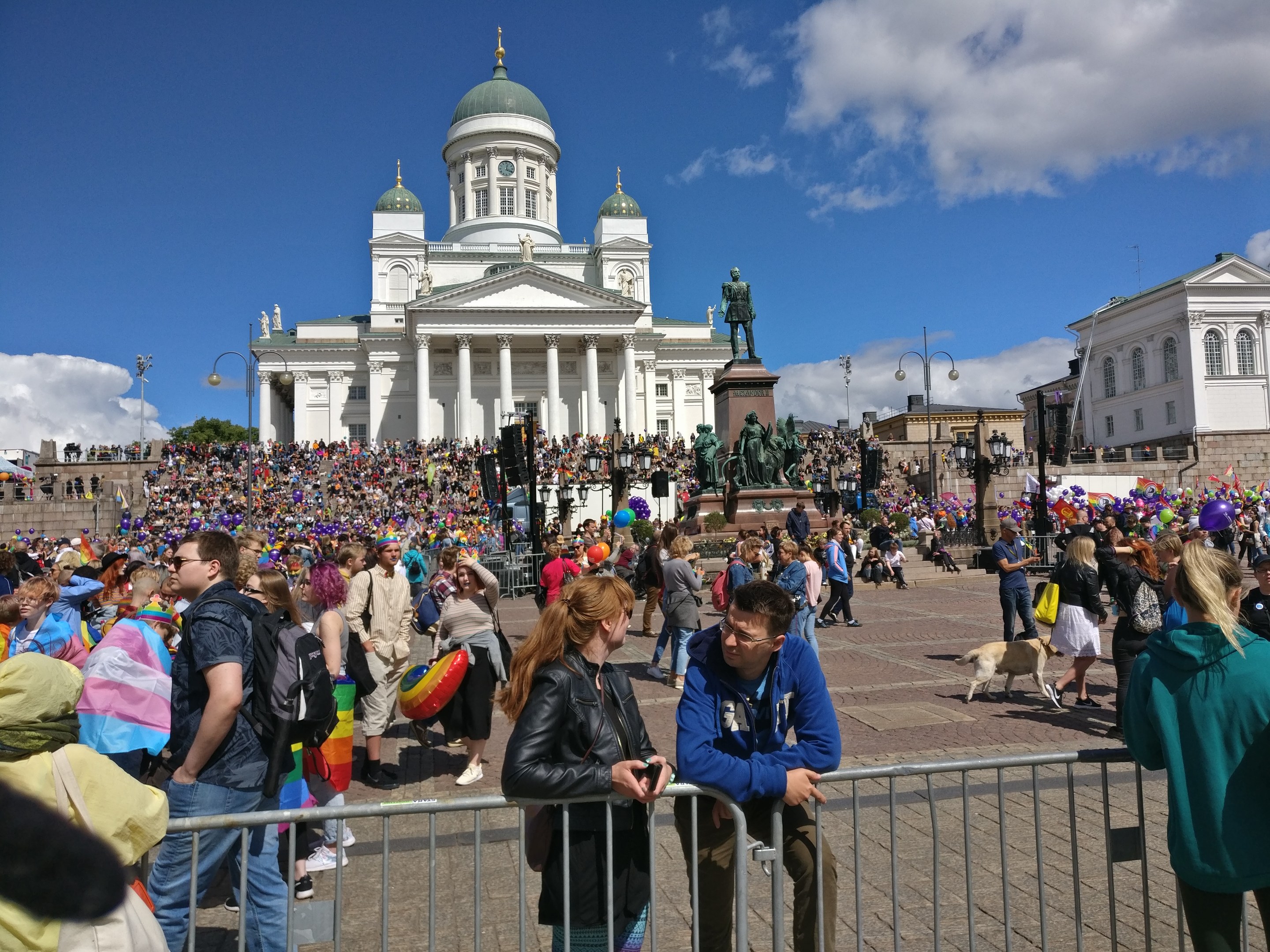 A large crowd stands outside a building with pillars, arches, and railings, surrounded by statues, trees, and light poles under a cloudy sky, many wearing bags and holding balloons during a Helsinki pride parade.
