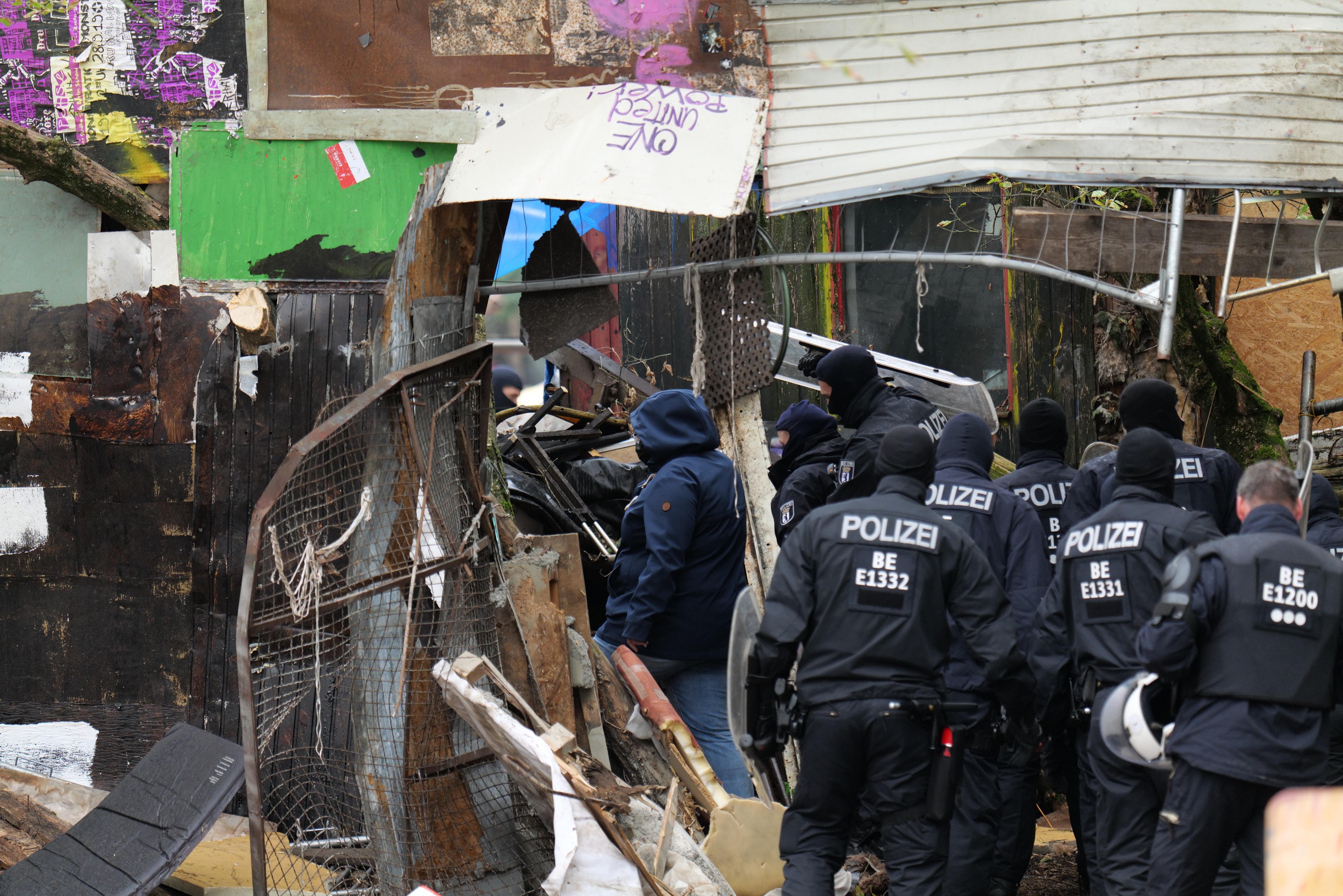Group of police officers in black uniforms and helmets standing in front of a destroyed building surrounded by debris and a metal fence.