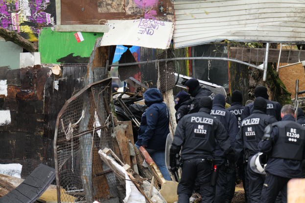 Group of police officers in black uniforms and helmets standing in front of a destroyed building surrounded by debris and a metal fence.
