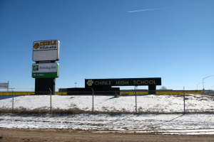 Eine Straße mit Schnee, ein Zaun mit Text, eine Wand, Laternenpfähle, Bäume und einen klaren Himmel, mit einem Schild im Vordergrund, das 'Chinle High School' lautet.