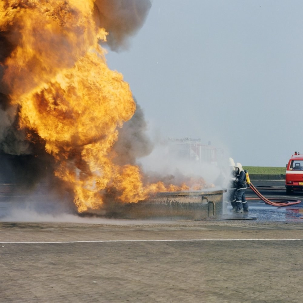 Feuerwehrfahrzeug steht in Flammen am Straßenrand mit zwei Helden, die Schläuche halten, ein Fahrzeug im Hintergrund und der Himmel.