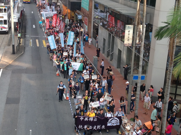 Ein großer Protestmarsch in Hong Kong mit Menschen, die Schilder und Plakate tragen, flankiert von Bäumen und Gebäuden auf der rechten Seite und Fahrzeugen auf der linken Seite.