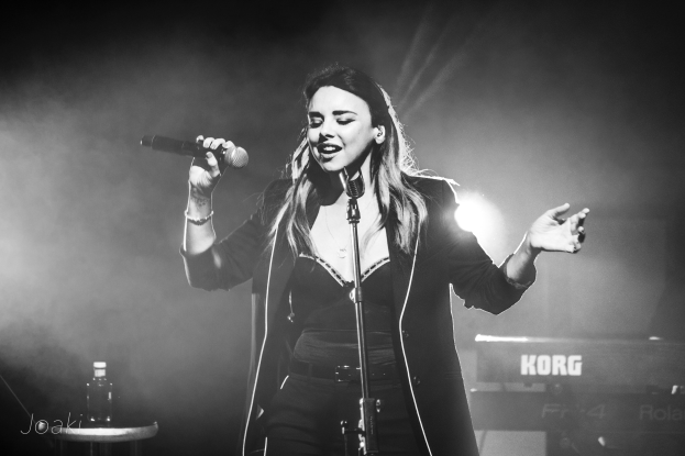 Demi Lovato performs on stage at the O2 Academy Brixton in London, standing at a microphone with a stand, with a table and bottle to her left, and musical instruments and stage lighting in the background.