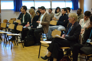 Mehrere Personen sitzen auf Stühlen in einem Raum, einige halten Gegenstände wie Laptops und Stifte, mit leeren Stühlen, Fenstern und Vorhängen im Hintergrund.