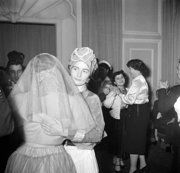 Schwarzes und weißes Foto einer Braut in einem Schleier und eines Bräutigams, der auf ihrer Hochzeitsfeier tanzt, umgeben von sitzenden und stehenden Gästen mit Vorhängen im Hintergrund.