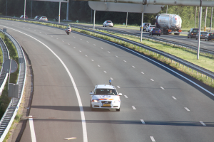 Ein Auto fährt auf einer Autobahn mit einem Polizeiwagen am Rande, anderen Fahrzeugen, Geländern, Pfosten, Planken und Bäumen im Hintergrund.