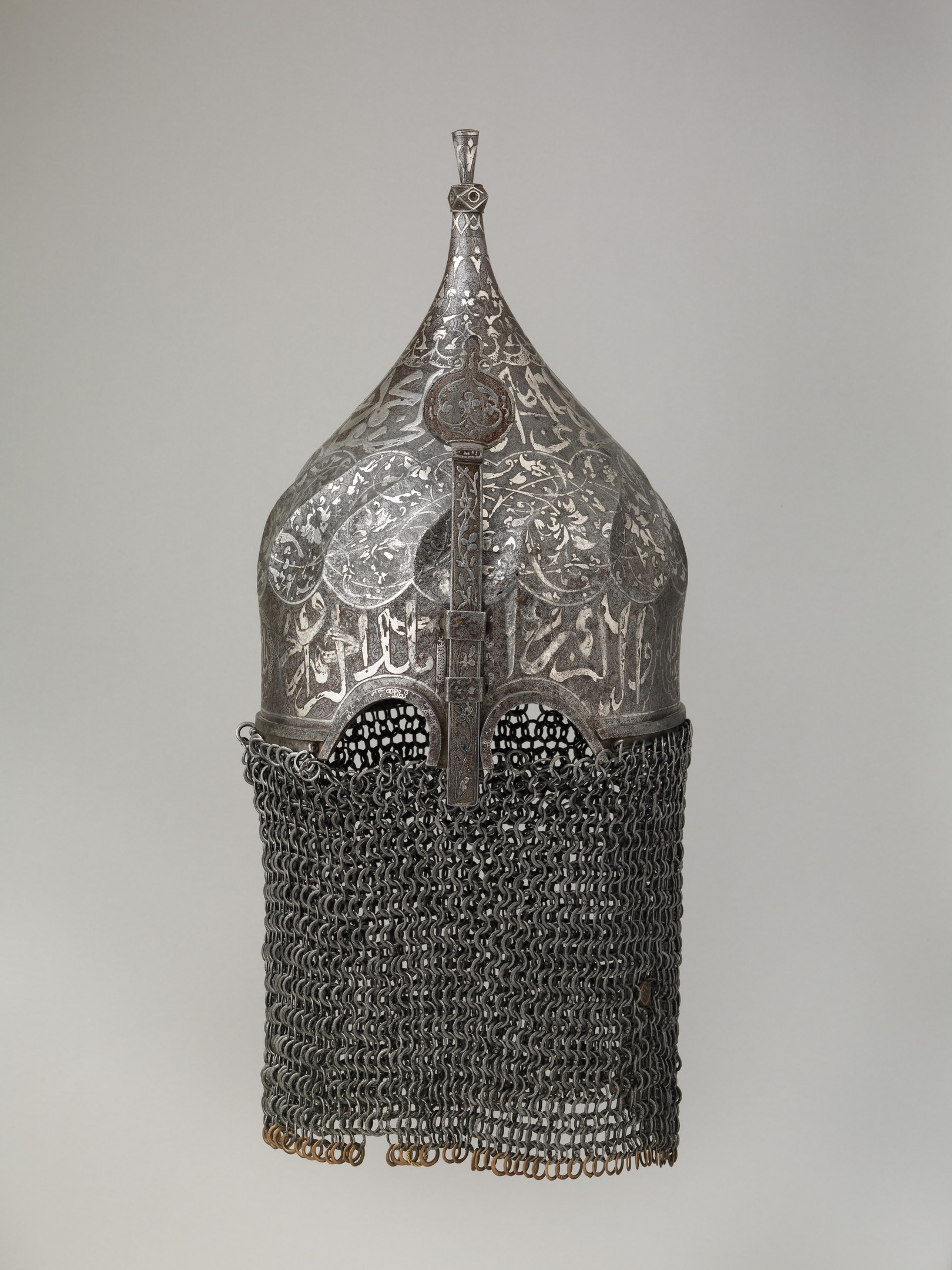 Close-up of a metal helmet with a chainmail design, featuring interlocking metal rings, set against a white background.