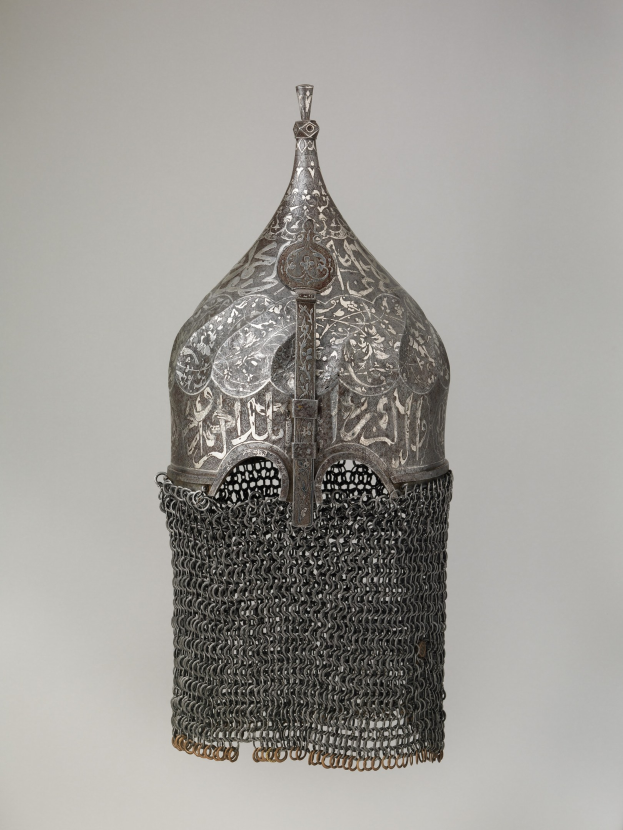 Close-up of a metal helmet with a chainmail design, featuring interlocking metal rings, set against a white background.