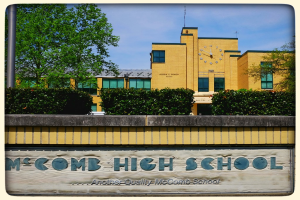 Ein Plakat mit dem Text "MCCOMB HIGH SCHOOL" auf einer Wand, mit Pflanzen, Bäumen, einem Pfahl, einem Gebäude und einem bewölkten Himmel im Hintergrund.