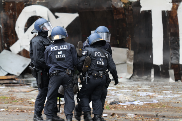 Polizeibeamte in schwarzen Uniformen und blauen Helmen stehen vor einem Gebäude mit Graffiti, umgeben von verstreuten Papieren und Gegenständen auf dem Boden.