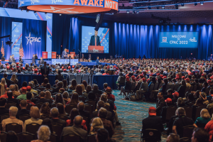 Eine große Menschenmenge sitzt auf Stühlen, die einer Bühne zugewandt sind, mit einigen Menschen auf dem Podium und auf der linken Seite, blaue Vorhänge und ein Bildschirm im Hintergrund und ein orangefarbenes Schild oben, das die CPAC-Veranstaltung am 25. Oktober anzeigt.