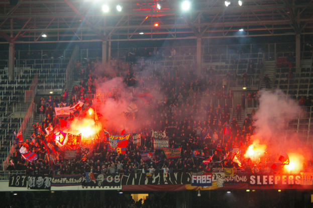 Eine große Menschenmenge in einem Stadion hält Fahnen und Banner hoch, während Raketen und Rauch aufsteigen und Metallrahmen und Deckenlampen über ihnen sichtbar sind.