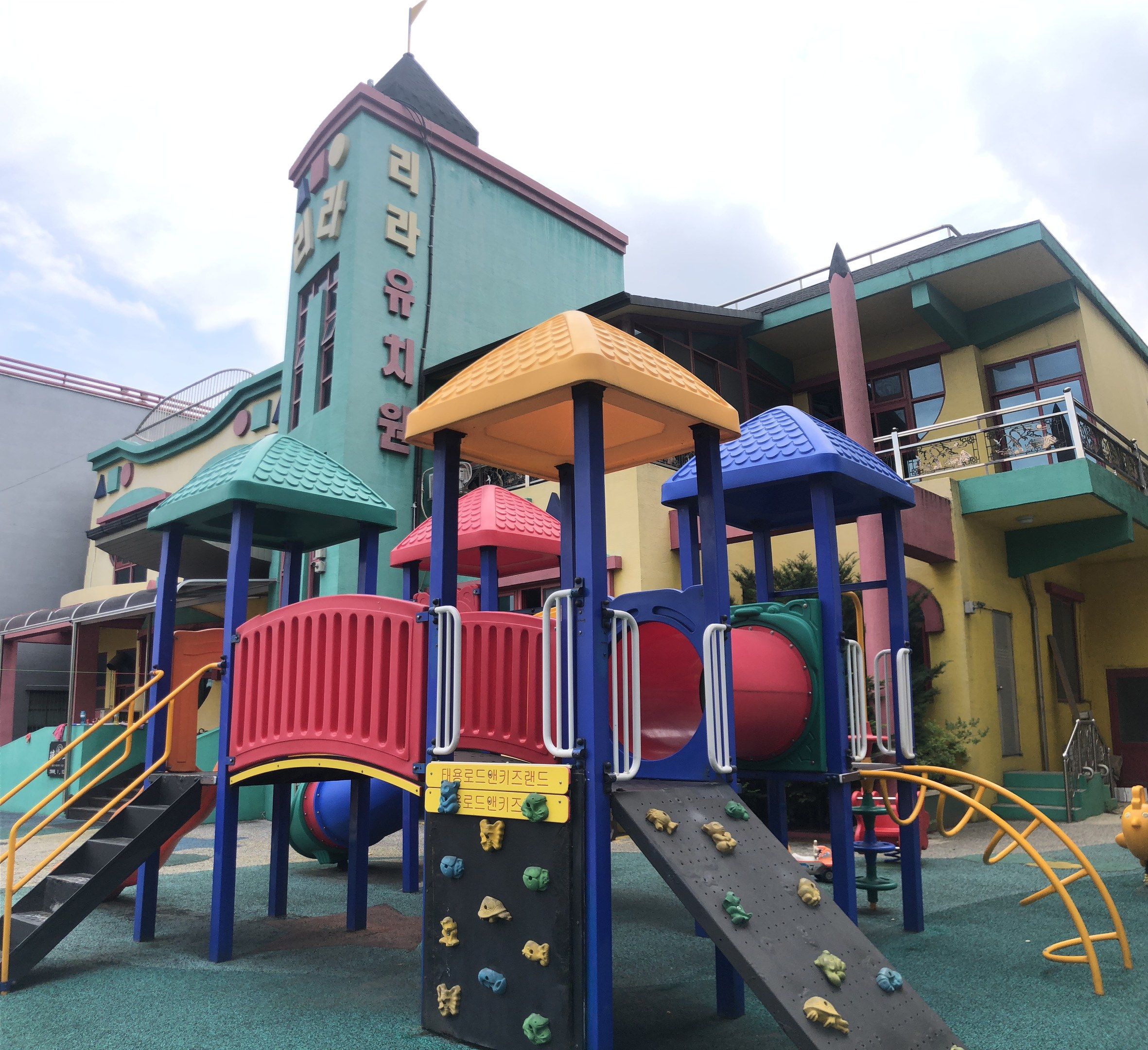 Bunte Kinderspielplatzgeräte zum Verkauf vor einem Gebäude mit Fenstern, umgeben von Bäumen und einem bewölkten Himmel.