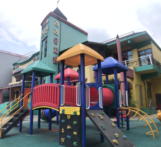 Bunte Kinderspielplatzgeräte zum Verkauf vor einem Gebäude mit Fenstern, umgeben von Bäumen und einem bewölkten Himmel.