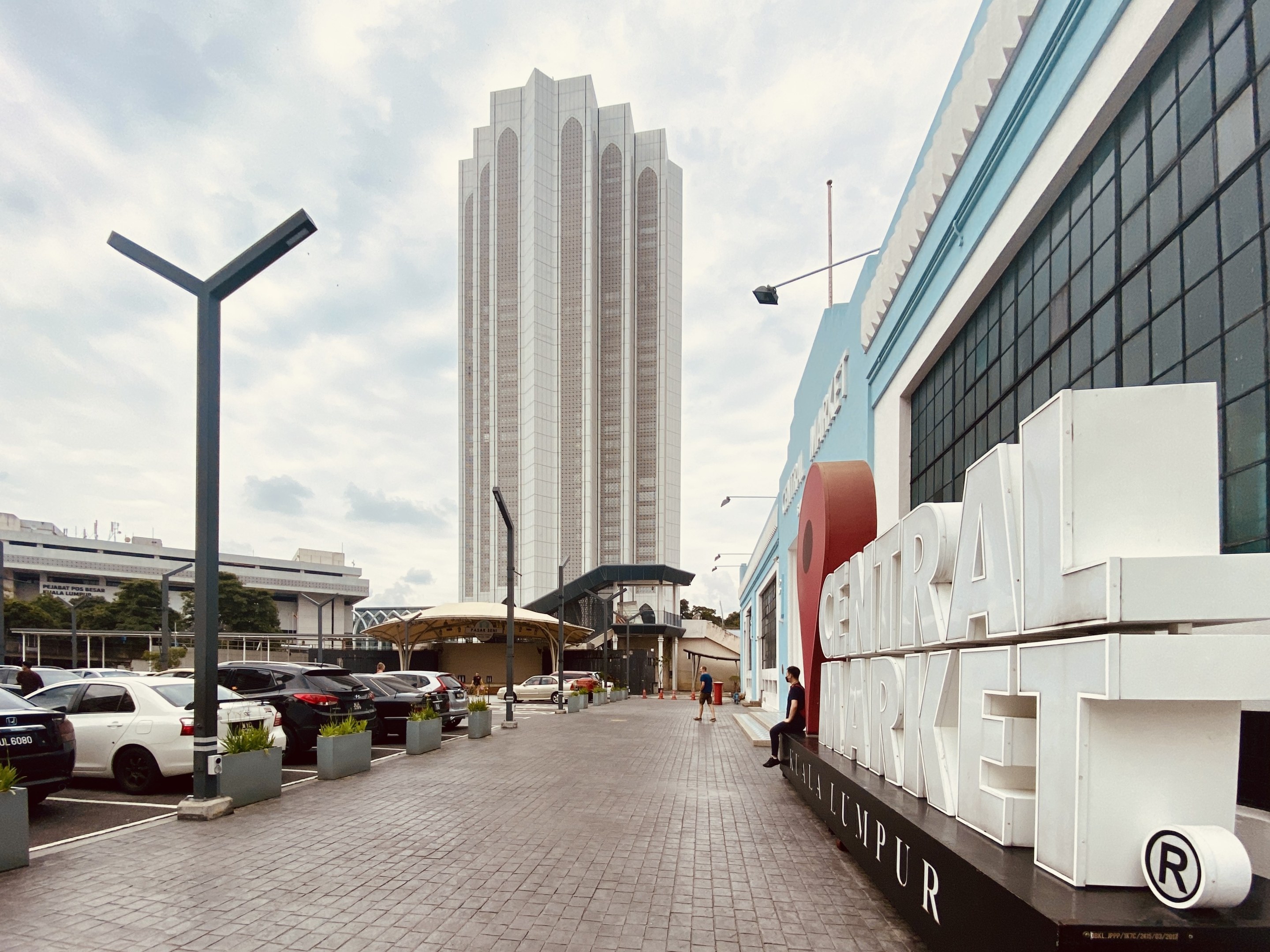 A busy central market in Kuala Lumpur, Malaysia, with tall buildings, vehicles, pedestrians, street furniture, and trees under a cloudy sky.