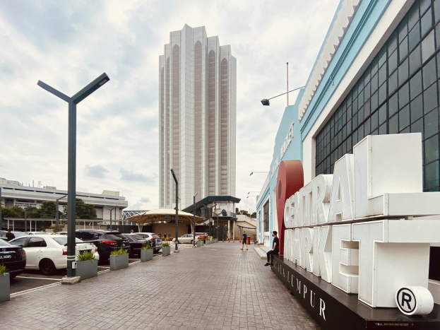A busy central market in Kuala Lumpur, Malaysia, with tall buildings, vehicles, pedestrians, street furniture, and trees under a cloudy sky.