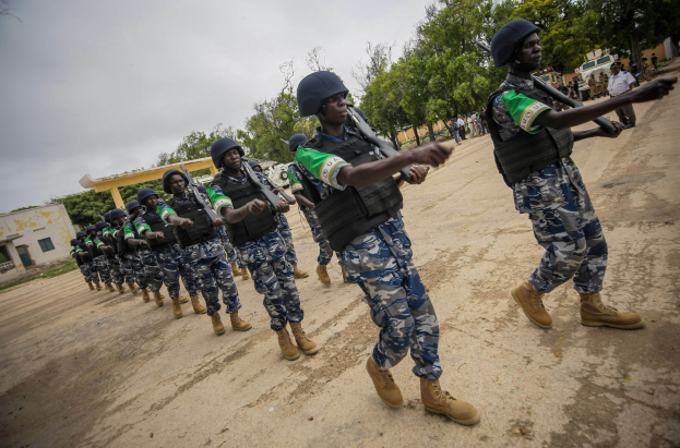 Nigerian soldiers marching in formation on a dirt road, with trees, buildings, and a vehicle visible in the background under a clear blue sky.