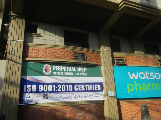 Außenansicht des Perpetual Help Medical Center - Las Pinas mit ISO 9001 2015-Zertifizierungsbanner, Säulen, Fenstern, Klimaanlagen und einem Pfahl mit einer Kette davor.