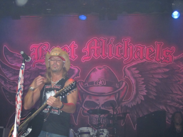 Ein musikalisches Konzert mit Scheinwerfern, einem Banner im Hintergrund, einer sitzenden Person in der rechten unteren Ecke, einem stehenden Gitarristen links mit einem Mikrofon davor und Gegenständen in der Mitte unten.