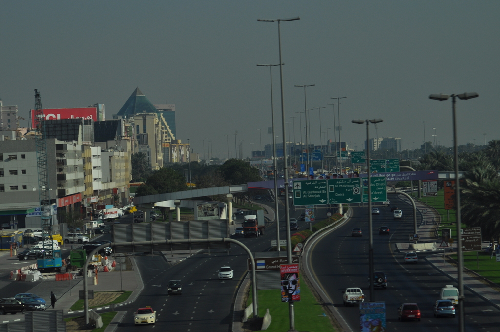 Eine Luftaufnahme einer Stadt mit Gebäuden, Türmen, Kraftfahrzeugen, Werbetafeln, Schildern, Straßen, Bäumen, Straßenlaternen, Menschen, Verkehrskegeln und Informationsanzeigen unter einem klaren Himmel.