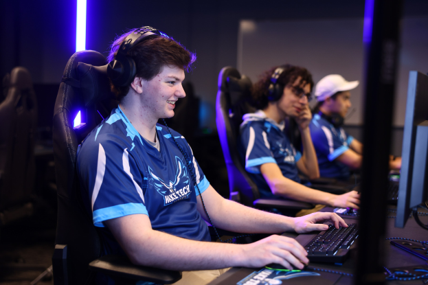 Eine Gruppe junger Männer mit Headsets lächelt, während sie an einem Schreibtisch mit Computermonitoren, Tastaturen und Mäusen sitzen, wahrscheinlich spielen sie ein Spiel als Teil eines Top-Esports-Teams.