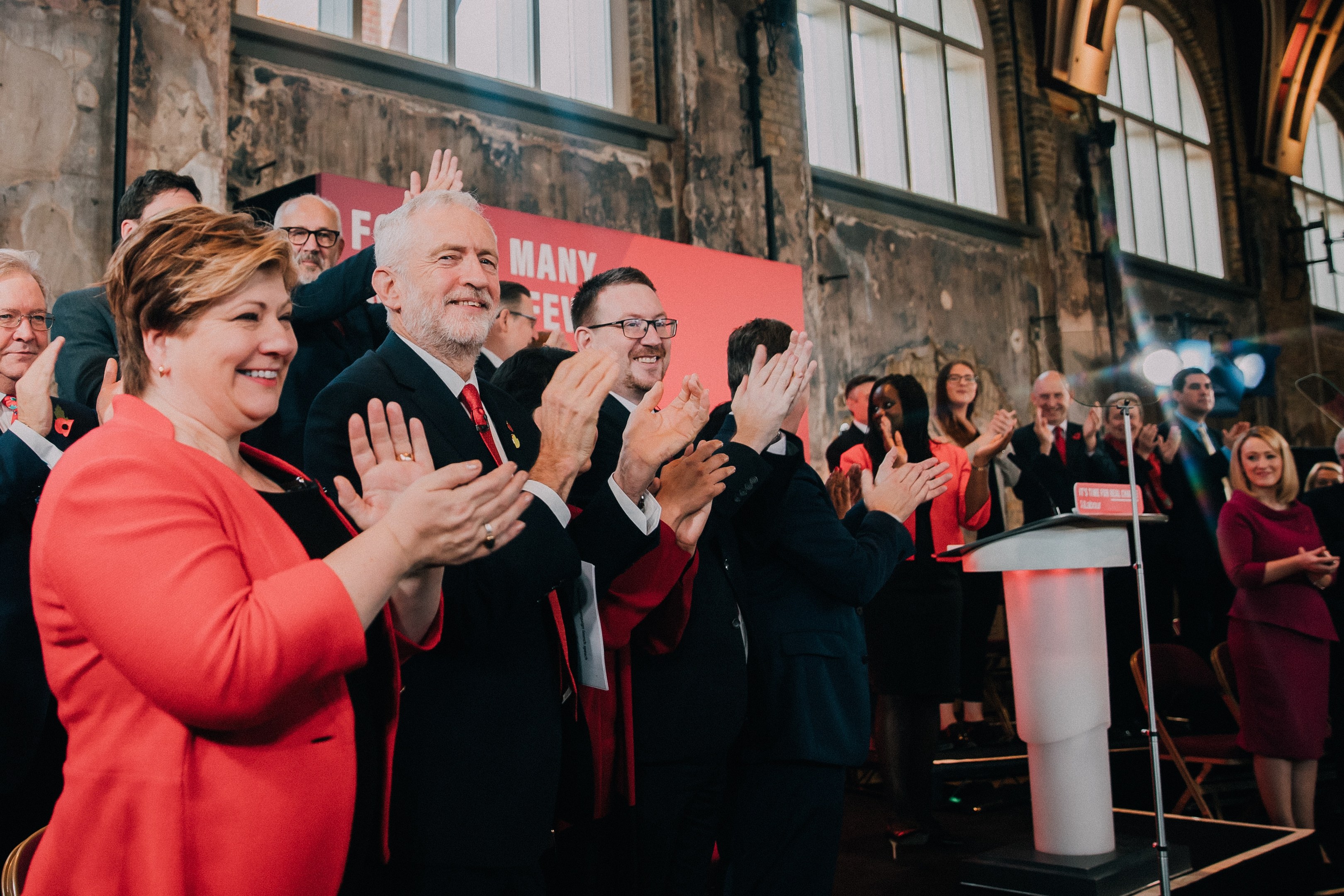 Eine Gruppe von Menschen, die vor einem Publikum applaudieren, mit einem Podium, Mikrofon und einer Tafel mit Text auf der rechten Seite und Stühlen, einer Fahne, einer Wand, Fenstern und Lichtern im Hintergrund.