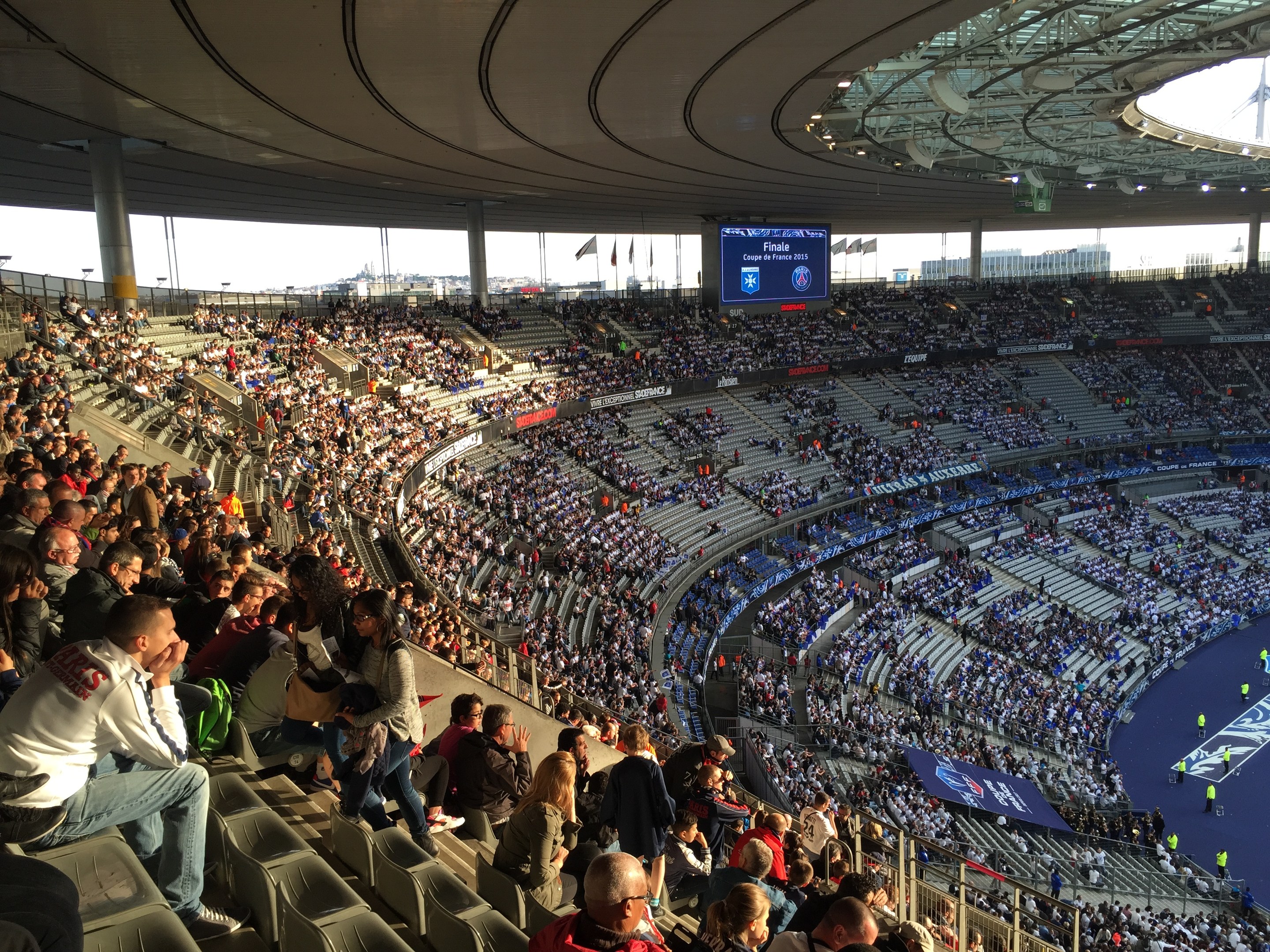 Large crowd in a stadium watching a soccer game with a stage, flags, poles, and a screen in the background, identified as the Allianz Arena in Munich, Germany.